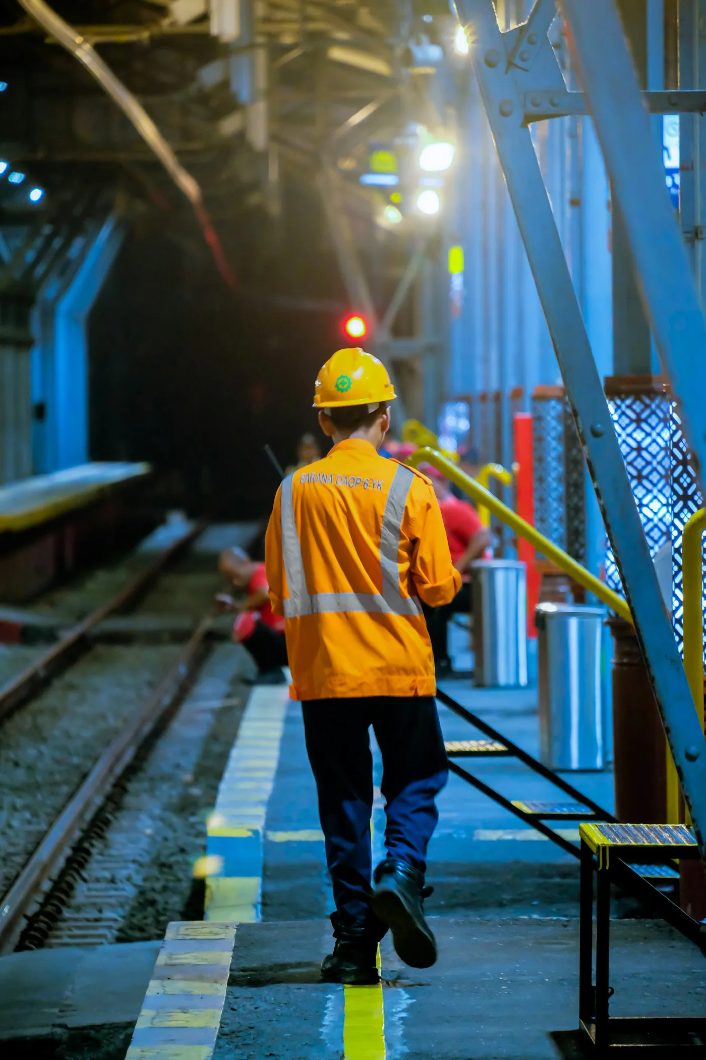A railway worker in an orange safety vest and hard hat walking a platform edge
